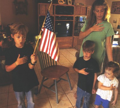 Photo: three home-schooled children and their mother hold their hands over their hearts and hold up a United States flag in their living room