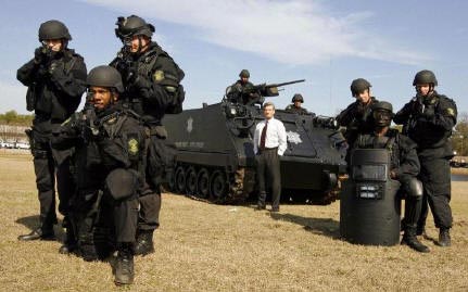 Here is a photograph of several men in dark uniforms with helmets and body armor, in combat posture, with assault rifles pointed directly at the camera. They are posed in front of a large tank. One man is standing in front of the tank, smiling, in a normal suit and tie.