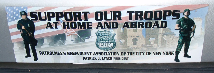 It features an armed, black-uniformed cop on the left and an armed soldier standing at attention on the right. In the background, a huge American flag flies over an image of a city building in New York and a ruined city presumably somewhere in Iraq or Afghanistan. Up to, it reads SUPPORT OUR TROOPS: AT HOME AND ABROAD, with a PBA shield and "Patrolman's Benevolent Association of the City of New York, Patrick J. Lynch, President," printed underneath.