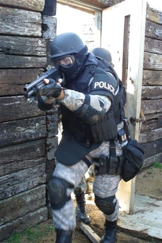 Here's a photo of a heavily armored soldier bursting through the door and pointing a shotgun at the camera. On his arm there's a band reading POLICE.