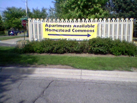 I have a photo of a large yellow banner draped over a fence on an island in the middle of the street, reading: “Apartments Available / Homestead Commons”