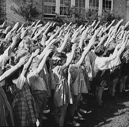 A crowd of white elementary school children with their right arms extended diagonally upwards.